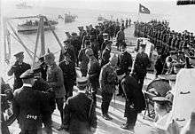 Several men in military uniforms, some wearing the Turkish Fez, aboard a warship; a large, boxy gun turret is on the right.