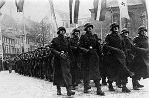 black and white photograph of helmeted soldiers marching through a town in the snow