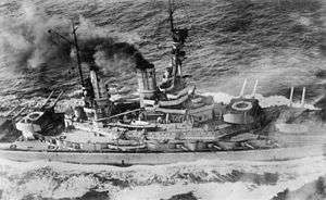 Overhead view of a large battleship; black smoke pours from its smoke stacks as it steams through choppy seas