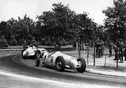 A front engined single-seated racing car leads a similar rear-engined car in a race.