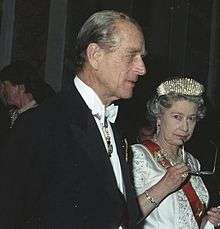 Elizabeth, in formal dress, holds a pair of spectacles to her mouth in a thoughtful pose