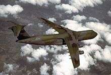 Four-engine jet transport with dark green and gray paint scheme in-flight above clouds.