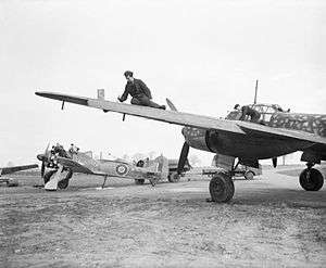 A photograph of a single–engined and a larger, twin engined aircraft on the ground. Two men are working on the wing of the larger aircraft. The engine–covers have been removed from the smaller aircraft and four men are working on its engine