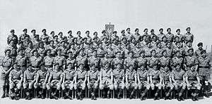 A formative black-and-white photograph of military personnel. The men wear khaki shirts and shorts with long, dark-coloured socks. They all wear dark berets.