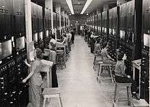 Two rows of control panels with dials and switches. Operators sit at them on four-legged stools