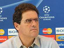 A man in a pale shirt, with curly brown hair. He is facing right and has narrow-rimmed glasses, and is in front of an advertising board