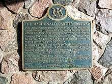 A blue plaque on a stone wall. The plaque has a yellow border, and is mostly rectangular in shape, with the long end oriented horizontally. However, the top side has a camel hump in the centre, with a circle centred at the top of the hump. Inside the circle is an Ontario coat-of-arms. The plaque reads: THE MACDONALD CARTIER FREEWAY This plaque commemorates the completion of the Macdonald-Cartier Freeway (Highway 401), the longest freeway operated without tolls by a single highway authority in North America. Covering 510 miles between Windsor on the United States border and the Ontario-Quebec boundary, it serves the richest economic region in Canada. In January 1965, it was named by The Honourable John P. Robarts, Prime Minister of Ontario, in honour of the two founding architects of the Confederation of Canada, Sir John A. Macdonald and Sir Georges Etienne Cartier. This site is located on the last section of construction, consisting of 15 miles between Ivy Lea and Highway 2, which was completed on October 11, 1968.