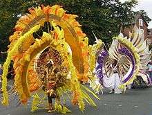 Part of a West Indian carnival procession passes along a public road in front of trees (to the left) and red-brick houses (to the right). In front is a participant wearing a gold helmet and vestigial armour. Above and around her is an enormous shield-shaped yellow, gold and red contraption supported by spokes and with many large feathers. Behind her is another participant in a white dress with a similar-shaped feathered shield contraption in yellow, brown and purple worn the other way up.