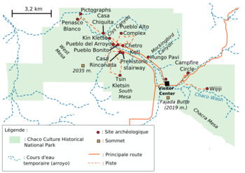 A large green area representing Chaco Culture National Historical Park's boundaries sits in the middle of a white field. The green area is roughly rectangular with one smaller square-like and one triangular appendage abutting it at bottom-left and bottom-right, respectively. Fifteen small red circles represent the location of important Chacoan sites; they are focused on a line running from top-left (northwest) to bottom-right (southeast). A dashed blue line depicting the Chaco Wash runs roughly along the same line; a network of dashed and solid orange lines represent trails and metalled roads, respectively, also focus on the same axis, connecting the red dots. Two gold squares define high points: "Fajada Butte (2019 m.)" and "West Mesa (2035 m.)".