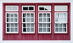 Four windows with white trim set in a red wooden wall. Each has a rectangular upper portion equally divided into six panes, projecting slightly over an identical bottom section. Above each is a smaller rectangular window with two panes