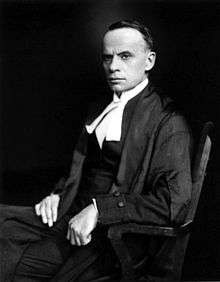 A dark-haired middle-aged man sits in a wooden chair, facing left. He is wearing judicial robes.