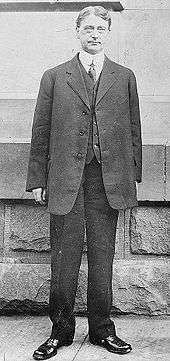 A middle-aged man in a suit and tie stands on a sidewalk in front of a stone wall.