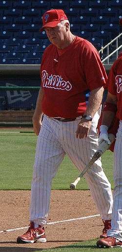 A man in a red baseball jersey, red baseball cap, and white baseball pants with red pinstripes walks on a baseball field.