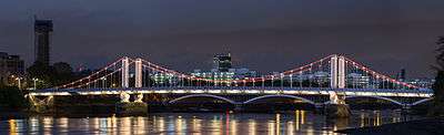 Night view of a lit suspension bridge over a wide river, which reflects the light from the bridge.