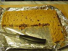 A foil-lined tray with a solid light brown sheet with dark brown spots. A section has been cut away and most of a knife is lying in that portion.