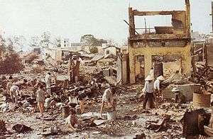 A number of Asian civilians standing amid ruined buildings and rubble.