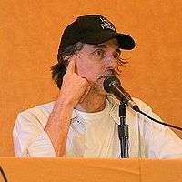 Upper body shot of man sitting behind desk with baseball cap speaking into a microphone