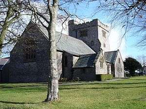 A low cruciform pale-coloured church seen from the southeast with the chancel prominent, beyond which is a broad castellated tower; in the foreground is the trunk of a tree standing in a grassed area