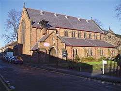 A church without tower or steeple seen from the side with clerestory; a porch and the aisle protrude; in front is a road with parked vehicles