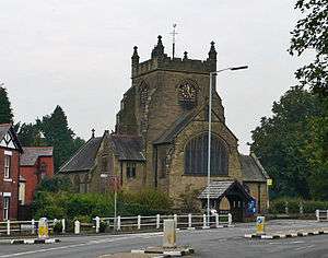 A cruciform church with a broad central tower; this is crenellated and has prominent buttresses; in front is a lych gate and a white railing; in the foreground is a lamppost and a road with traffic islands