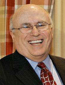 A bald bespectacled man wearing a pin-striped suit, a blue shirt, and a red tie laughs. Behind him is a red, brown, and white patterned drapery