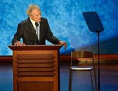 Man in coat and tie stands at a podium, looking towards a chair.