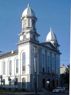 A formal building with arched windows and two domed towers is on a street corner near other less formal buildings. A car is parked in front of the formal building. Diagonally opposite are a stop sign and a small stone sculpture or monument.