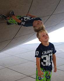 A boy stands vertically with his head touching the mirror-like surface of an object; his reflection in the object appears horizontal