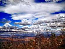 View from a high vantage point of a mountainous landscape beneath a party cloudy sky, there are twisted small trees in the foreground