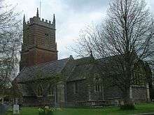 Stone building, partially obscured by trees. Red brick tower with horizontal stripe pattern surmounted by battlements.