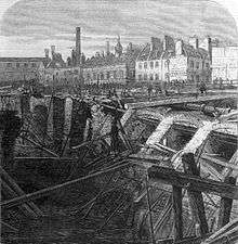 drawing showing a large quantity of collapsed brickwork retaining wall and destroyed timber scaffolding alongside a flooded excavation. Beyond the cutting is a wide open area with small figures inspecting the damage. The backdrop is formed by a collection of two storey buildings many of which have large chimneys.