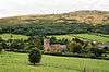 Church building with square tower seen within green fields with trees and hills behind.