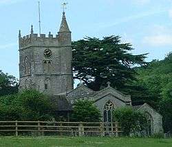 Gray stone building with arched windows. Square tower topped with spirelet, flagpole and weather vane. Foreground has small trees and bushes and a wooden rail fence.