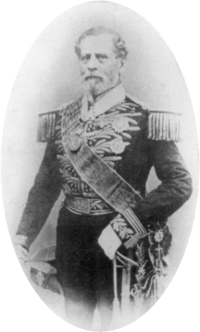 Tintype photograph depicting a full-length portrait of an older, bearded man wearing an elaborately embroidered military dress uniform with heavy epaulets, sash of office, sword, gloves and holding a bicorn hat in his right hand