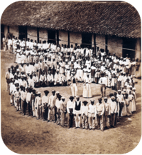 Photograph showing a group of people dressed in white, who have gathered in front of a tile-roofed farm building and observe another large group which has formed a large circle surrounding 5 men straddling large drums, a woman and 2 other men.