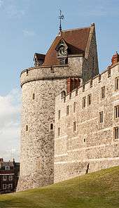 A photograph of a castle tower, the tower is pierced by small windows and has a coned, red-tiled roof, with a clock built into one side. The sky behind the wall is pale blue.