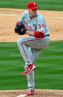 A man in a gray baseball uniform with red trim and a red baseball cap holds an unseen baseball with his right hand while wearing a black baseball glove on his left; he has one leg lifted partially in the air