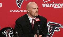 Color head-and-shoulders photograph of bald white man with reddish goatee (Dan Quinn), wearing navy suit jacket, white dress shirt and red-striped necktie, seated at a press conference table in front of a red Atlanta Falcons backdrop.