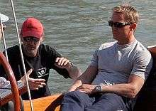 A man wearing a grey shirt, jeans and sunglasses sits on a boat at sea. Besides him, a man wearing a black shirt, red cap and sunglasses talks to another which is mostly off the picture.