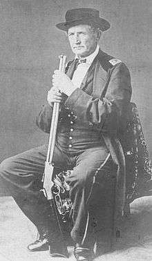 An older man wearing Civil War-era military garb and a hat sits on a chair and holds a lever action rifle.