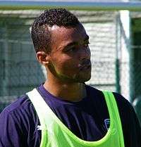 A photograph of a black man with short black hair wearing a light green vest over a navy blue shirt.