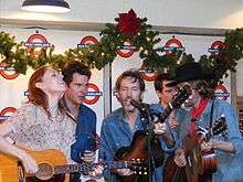 A group of musicicans, including Welch and four men, crowd around two microphones, passionately singing. There are three guitarists and one man playing a stand-up bass.