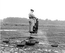 An Army officer in khakis and cap stands aboard a strange flying machine with twin rotors beneath his feet, running up on the ground, performing pre-flight checks.