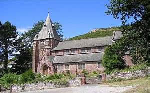 A church seen from the south with the edge of a hillside beyond; it has a clerestory and, to the left a tower with a short spire; in front is a wall with two gates and the whole is framed by trees on each side