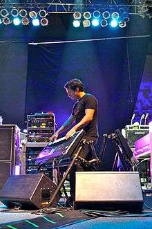 Derek Sherinian on stage playing a keyboard mounted on an angled keyboard stand. In the background are amplifiers, effects racks, a guitar rack and stage lighting mounted on the roof.