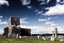 A ruined stone building with ecclesiastical stylings, surrounded by short grass and scattered headstones, with a stone round tower to the right. In the background, a stretch of calm water can be seen in front of a tree-lined shore.