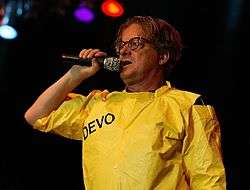 Mothersbaugh holding a microphone to his mouth, wearing a bright yellow costume