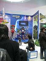 Inside a large, brightly lit convention center room with white walls is positioned a promotional display booth for a video game. A saleswoman clad in a blue shirt and skirt and a red bowtie motions towards several illustrations on the booth, explaining their implications. The illustrations are anime-styled and depict several outlandish and brightly colored creatures. Three men in dark jackets watch the demonstration.