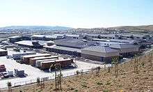 Several school buildings, nearly finished but with exposed cement walls where stucco will be. The sidewalk and road in front are also incomplete, and gravel is found in their place. The parking lot in front is full of trailers and trucks used by construction personnel.