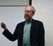 A grey-haired man with a grey beard wearing black jacket and grey shirt, talking with a whiteboard behind him.